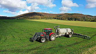 Ein roter Traktor fährt mit einem Güllefass mit Schleppschlauch-Verteilung über ein junges Getreidefeld und bringt Gülle aus.