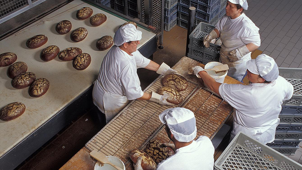 Vier Personen in weißer Wäsche arbeiten in einer Bäckerei. Auf einer Ablage liegen knusprige Brote.