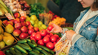 Junge Frau mit Einkaufsnetz vor Obst- und Gemüseregal greift nach einer Tomate. Junge Frau mit Einkaufsnetz vor Obst- und Gemüseregal greift nach einer Tomate.