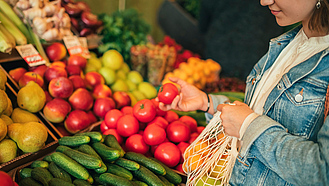 Junge Frau mit Einkaufsnetz vor Obst- und Gemüseregal greift nach einer Tomate.